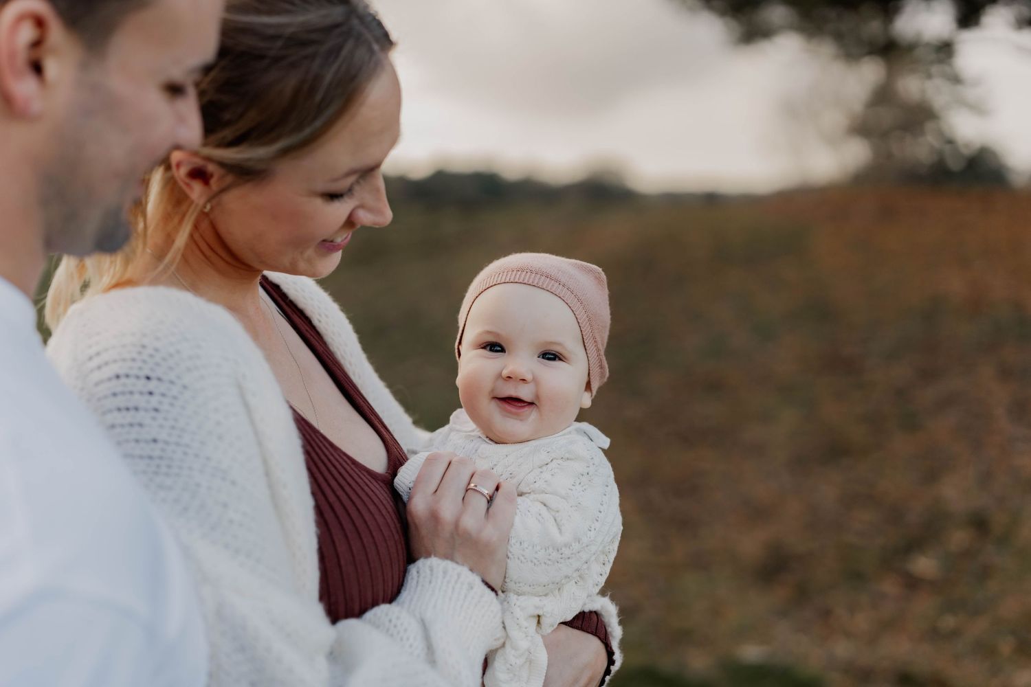 Family portrait with baby in white knit clothing against natural outdoor backdrop.