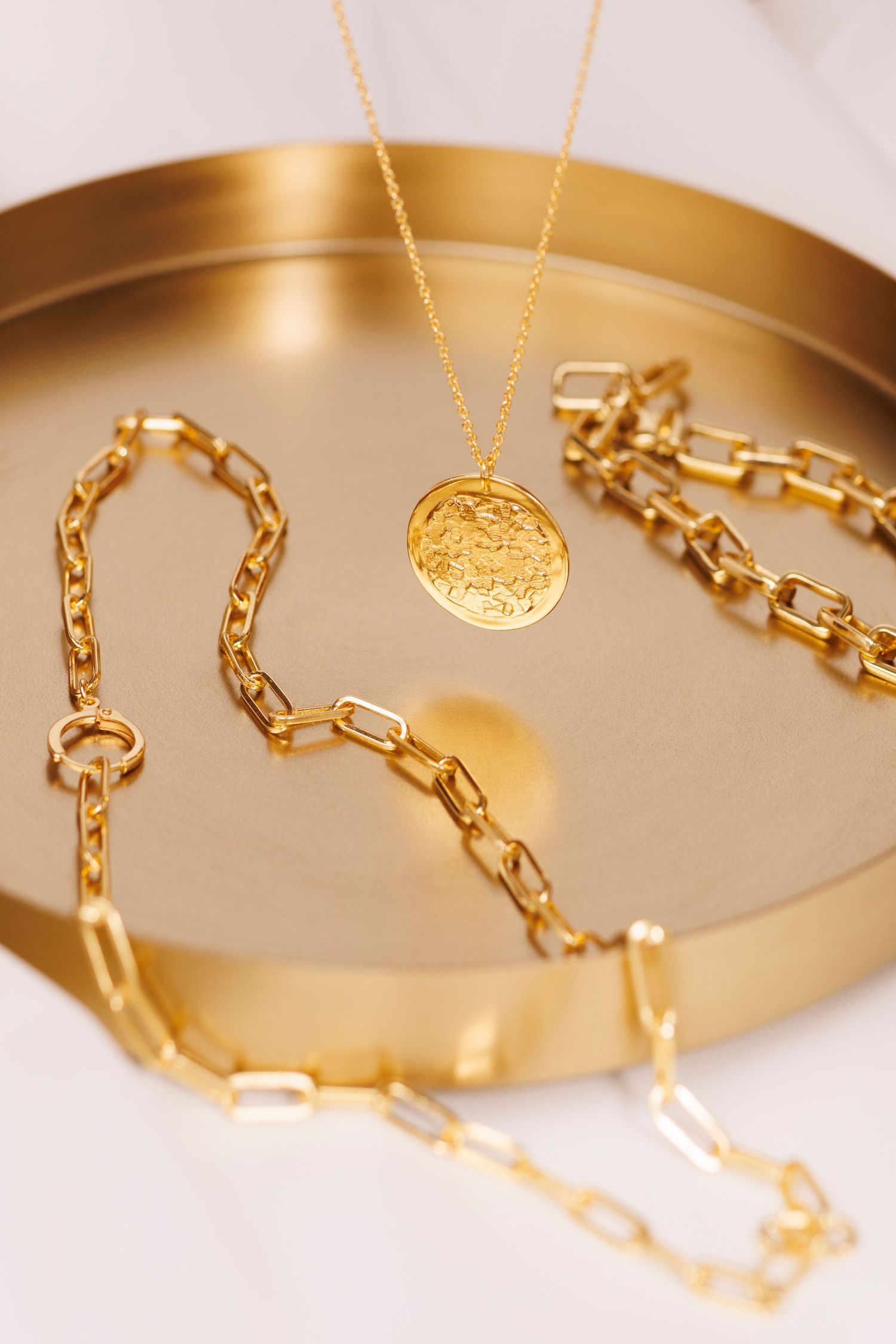 Gold chain necklace and medallion pendant displayed on metallic gold tray surface.
