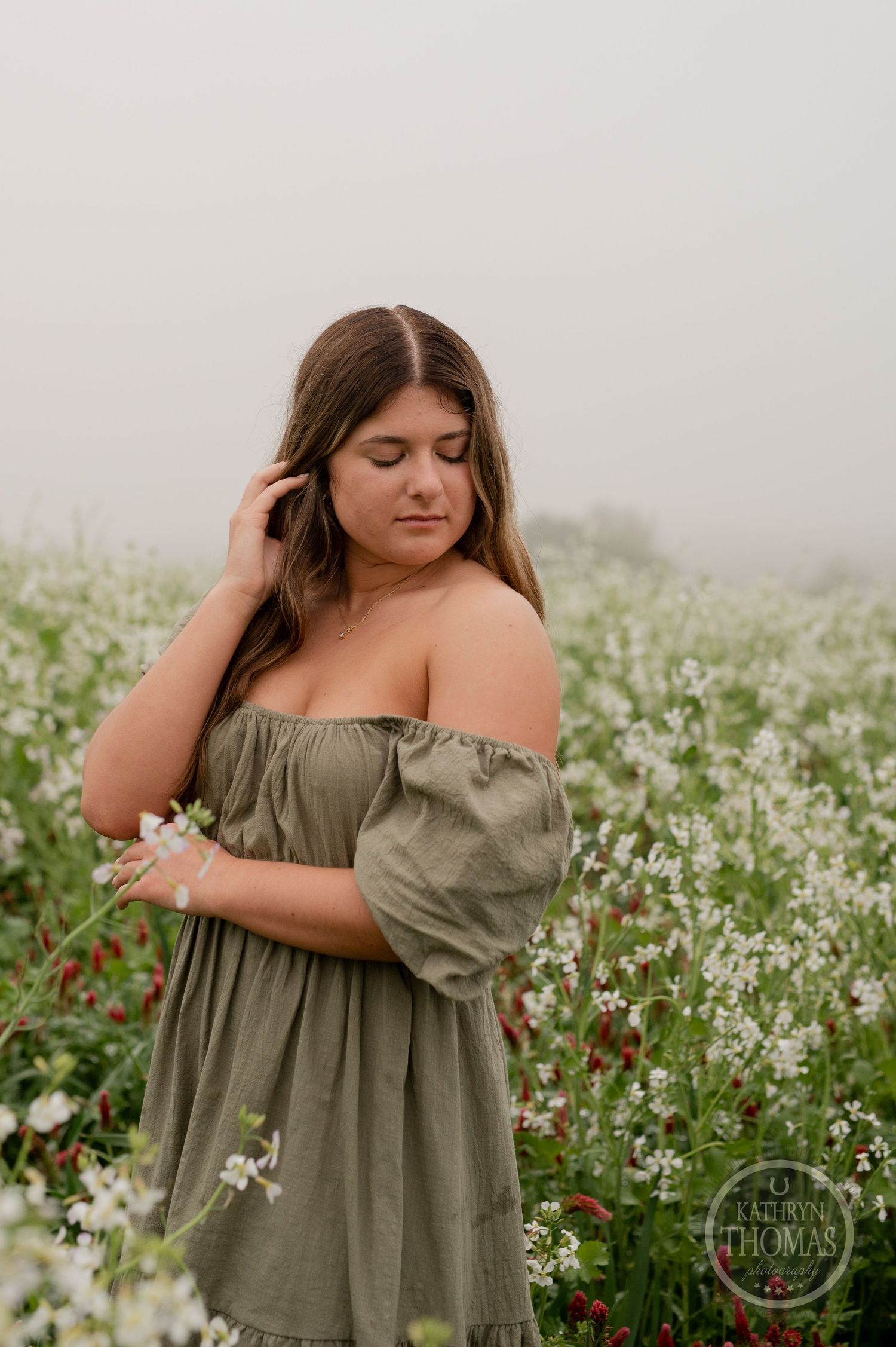 Model in olive dress looks down thoughtfully while standing in a flowering meadow.