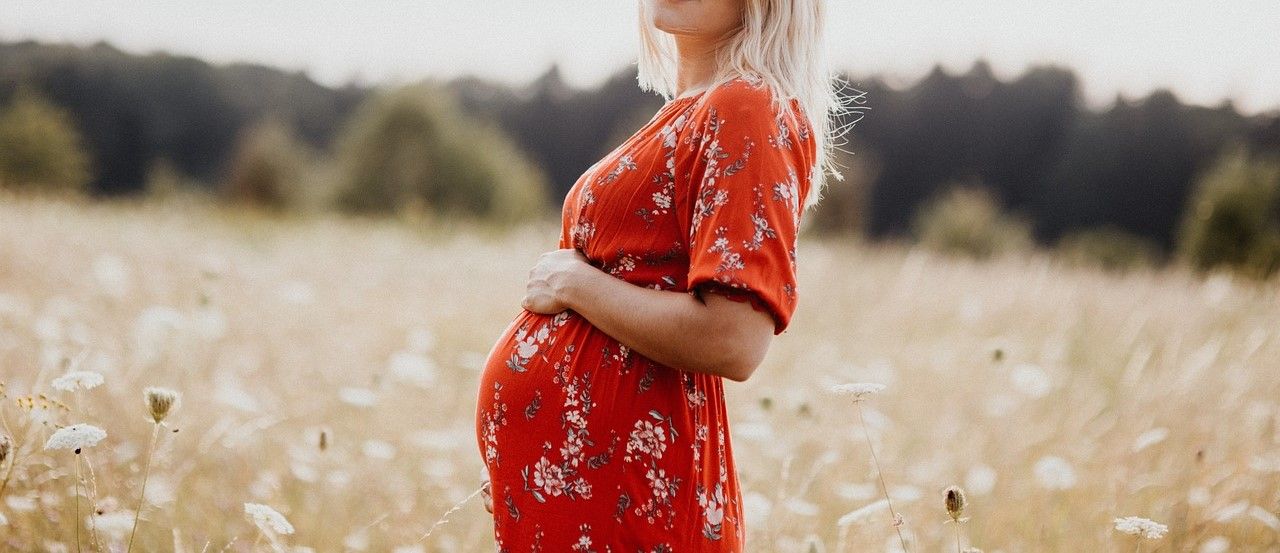 Kvinna i röd klänning som står på ett fält. Hon är gravid och håller ena handen på sin runda mage.