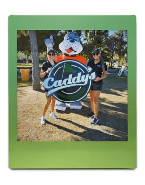 Two people (mom and daughter) in athletic wear pose outdoors with a large circular Caddy's logo sign and mascot in a park setting.