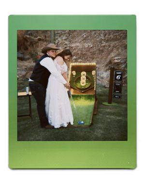 A bride and groom at a weeding in formal attire posing next to a Skeeball game setup at an outdoor event with a green target board.