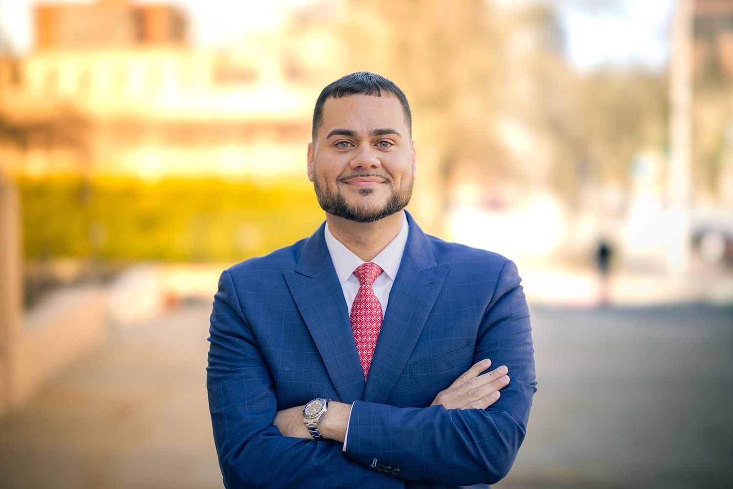 Professional in a navy suit and coral tie poses confidently with crossed arms against a blurred outdoor background.