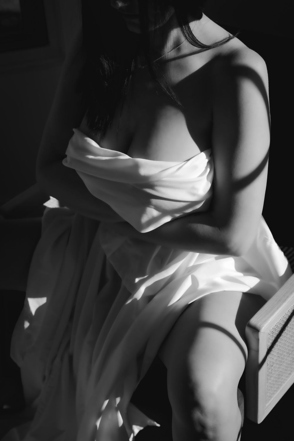 Black and white boudoir image of a nude woman covered in a white sheet with dramatic lighting flowing in from a nearby window.