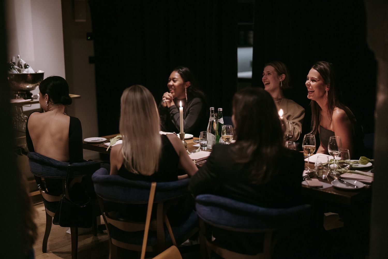 Intimate dinner party scene with guests gathered around a dimly lit table in a cozy restaurant setting.
