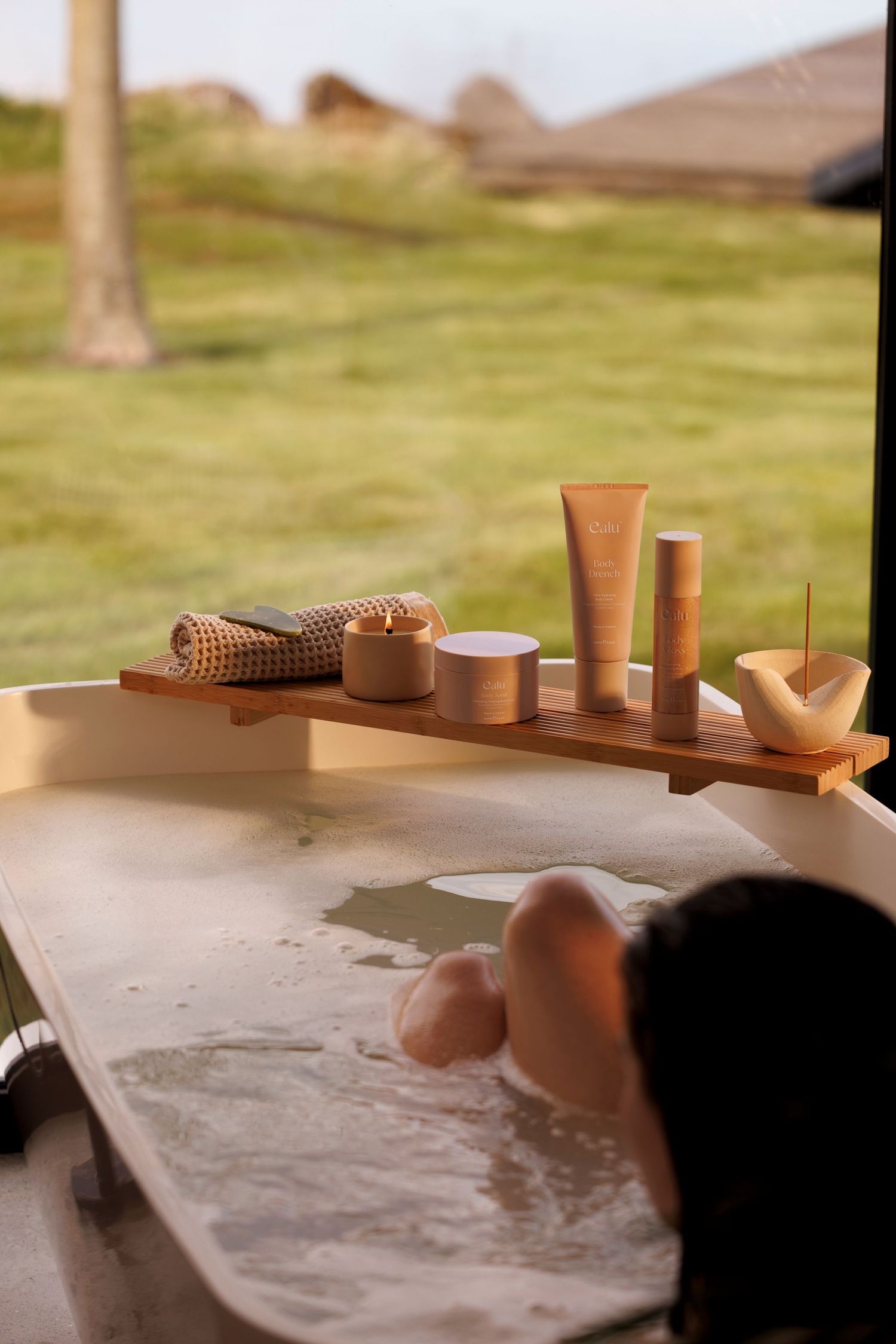 Luxurious bath setup with organic beauty products displayed on wooden tray while someone relaxes in bathtub.