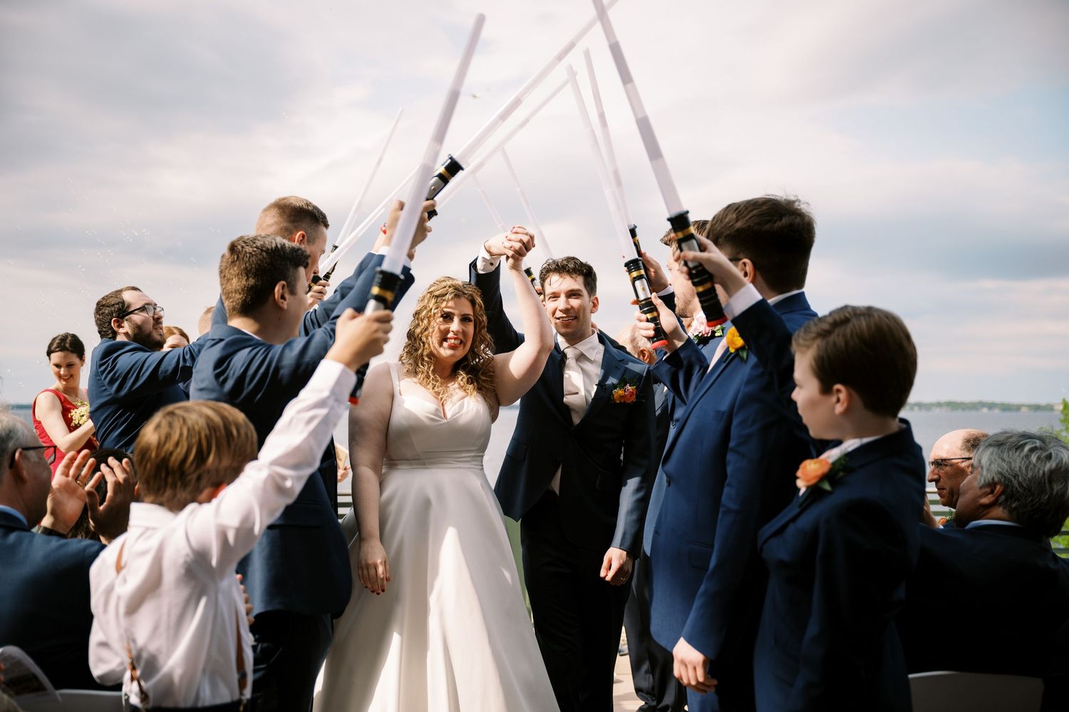 A wedding celebration with guests raising their arms in a celebratory archway against a cloudy sky backdrop.