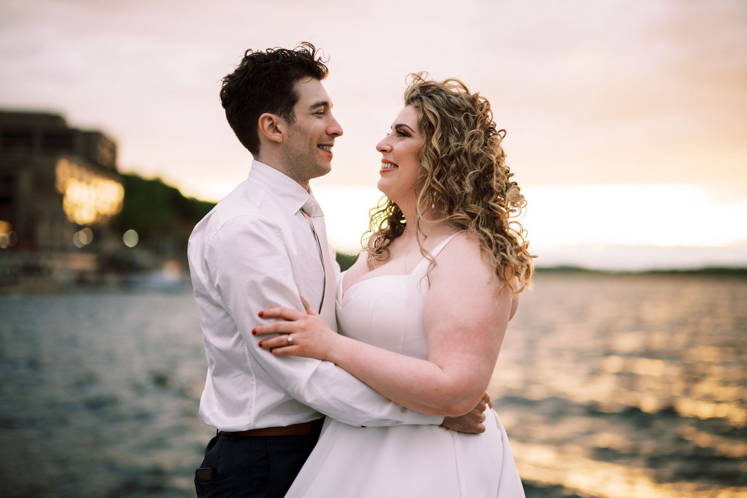 A happy couple embraces by the water during sunset with sparkling waves and warm lighting in the background.