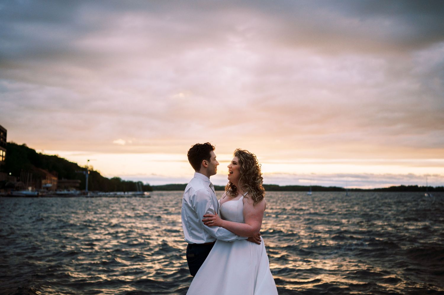 A couple embraces near the waterfront at sunset with rippling water and coastal scenery in the background.