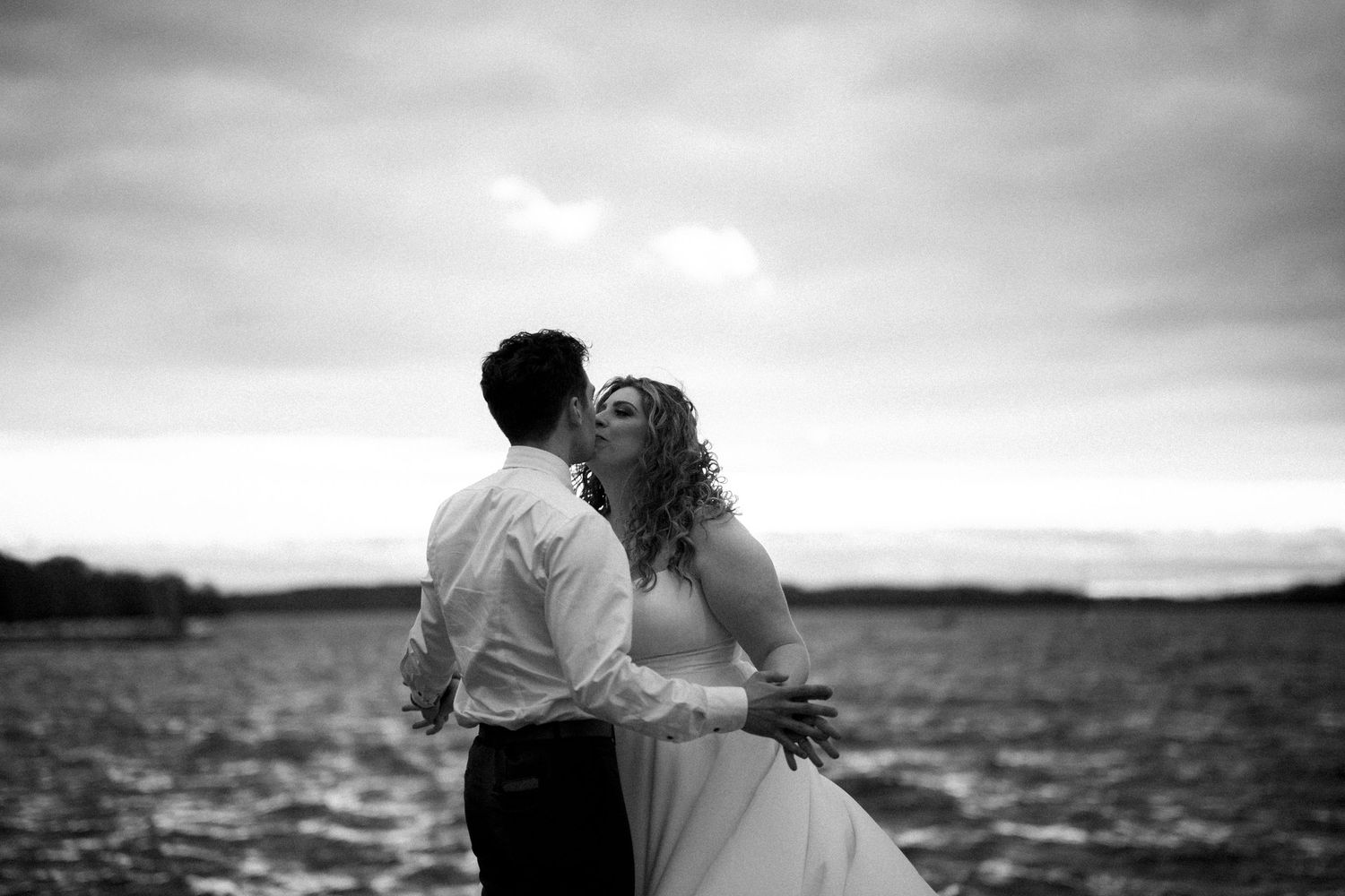 Black and white romantic portrait of a couple overlooking the water on their wedding day against a dramatic cloudy sky.