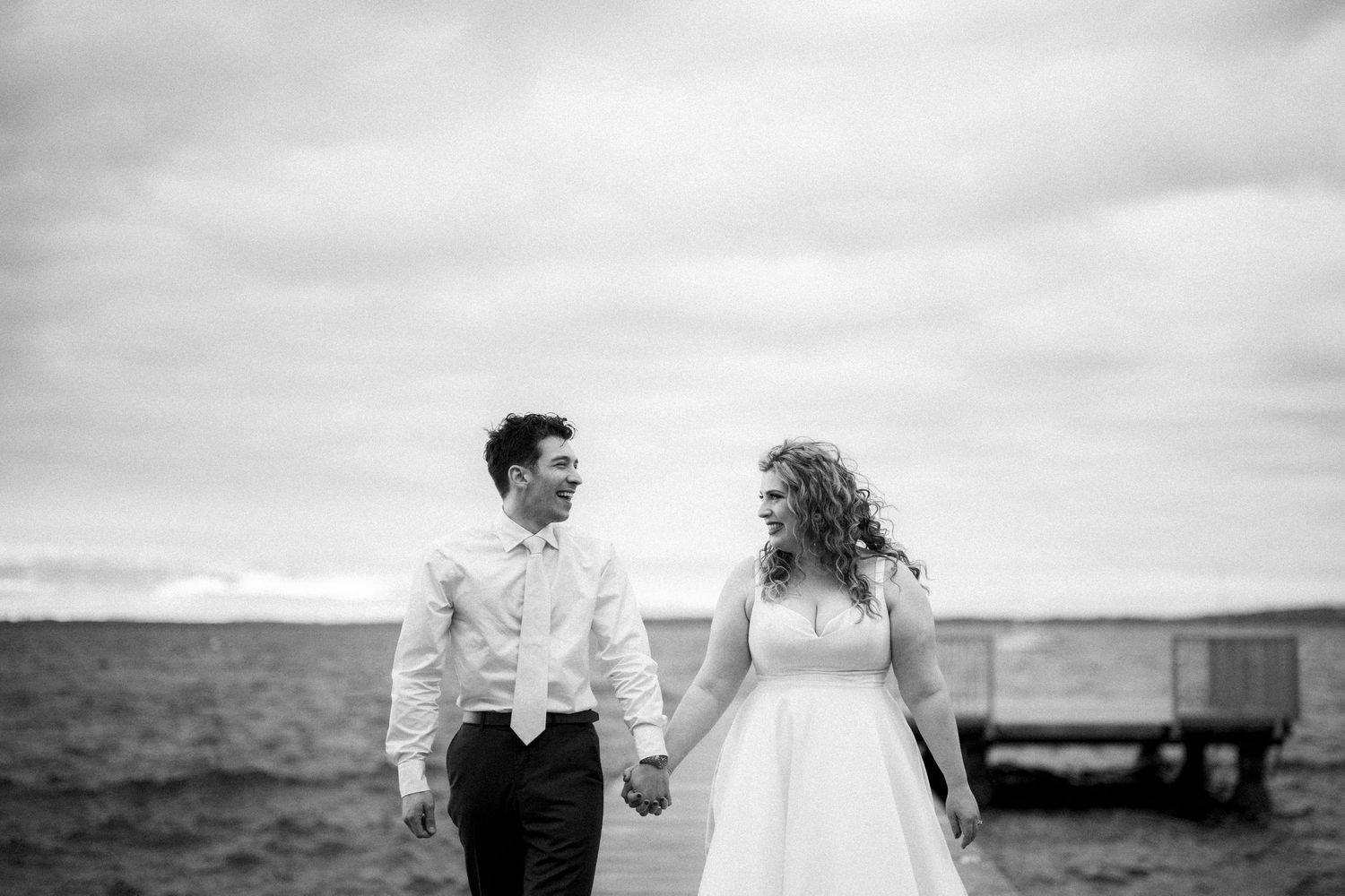 A couple on their wedding day shares a romantic moment while walking along a dock by the ocean in black and white.