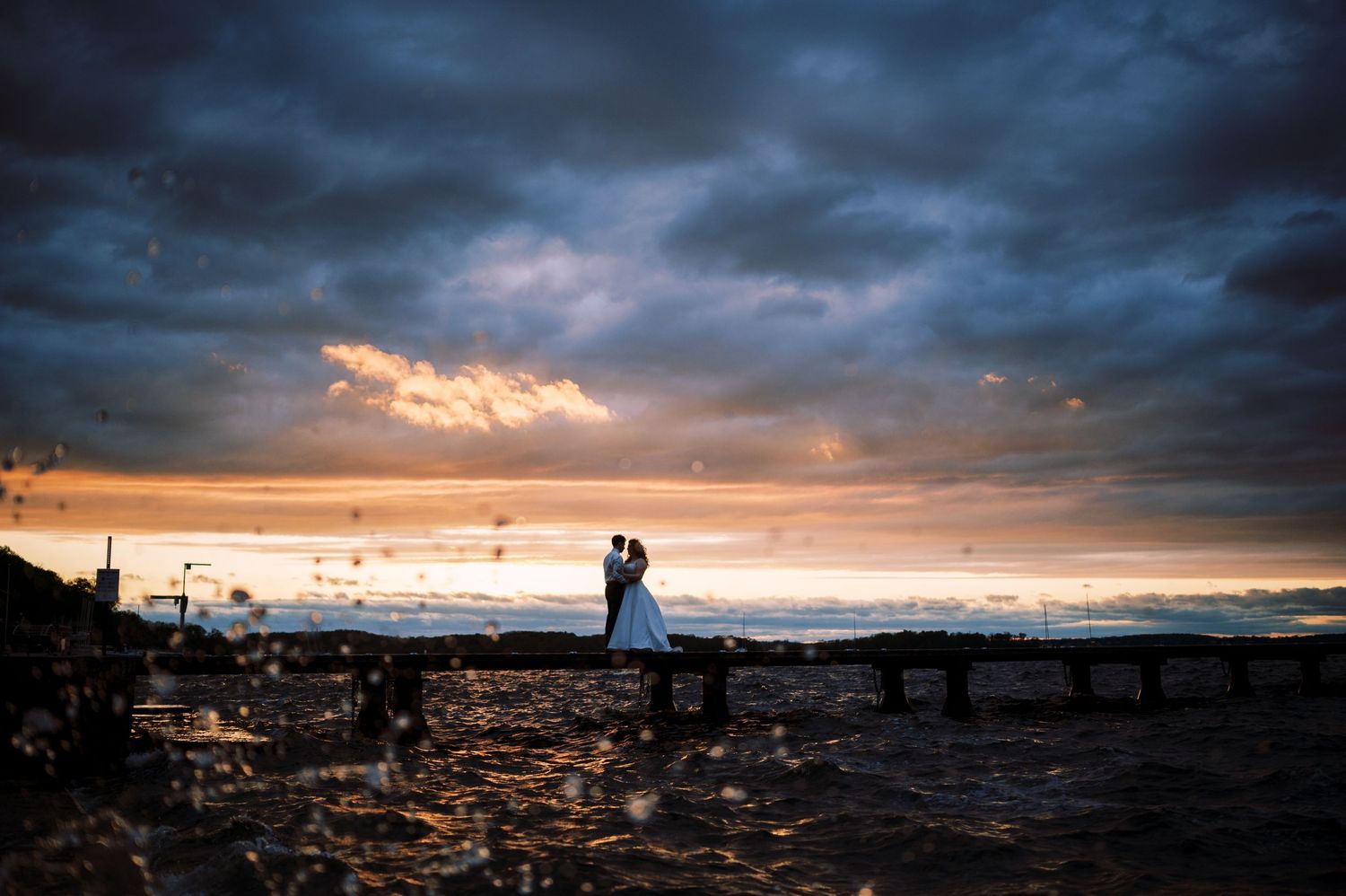Two silhouettes stand together on a beach at sunset with dramatic storm clouds and golden light over the ocean.