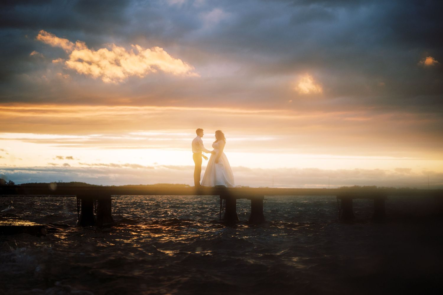 Two silhouettes stand together on a hilltop against a dramatic golden sunset sky with glowing clouds.