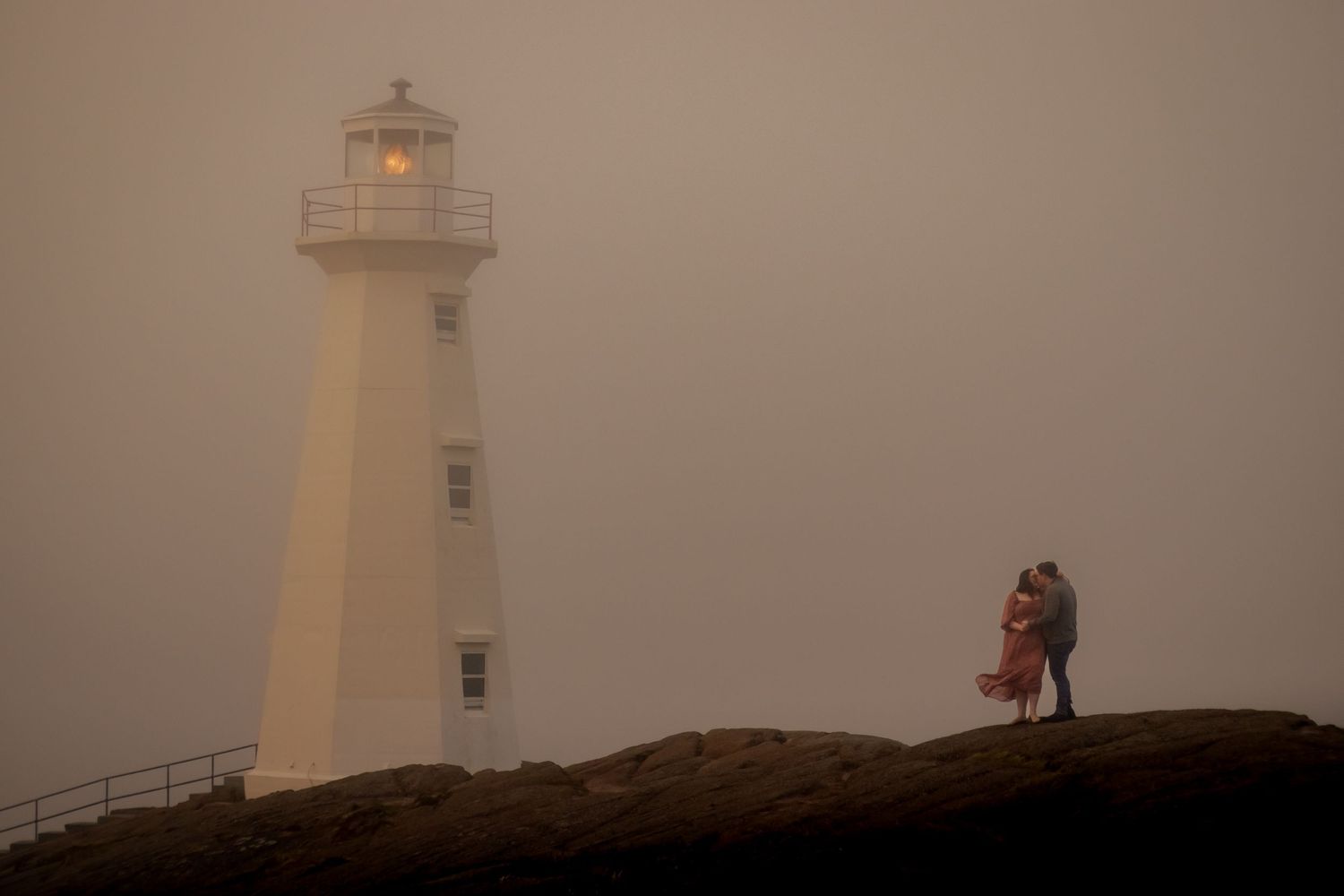Two silhouettes embrace near a lighthouse on a foggy, atmospheric evening at Cape Spear in Newfoundland.