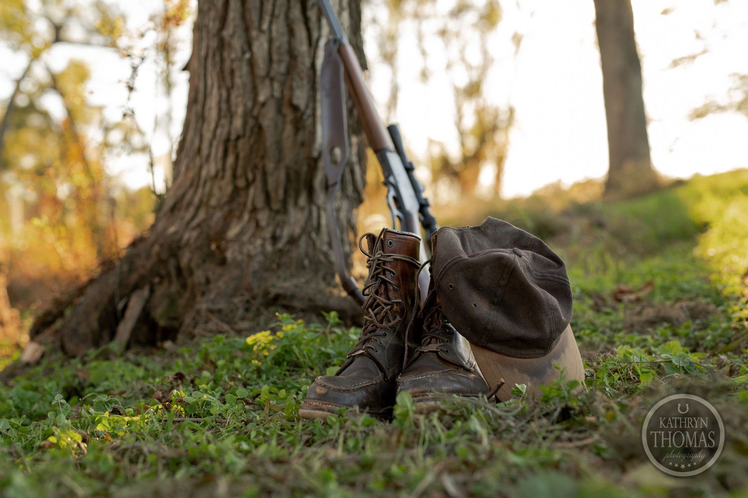 Hunting boots and rifle resting against a tree trunk in dappled sunlight.