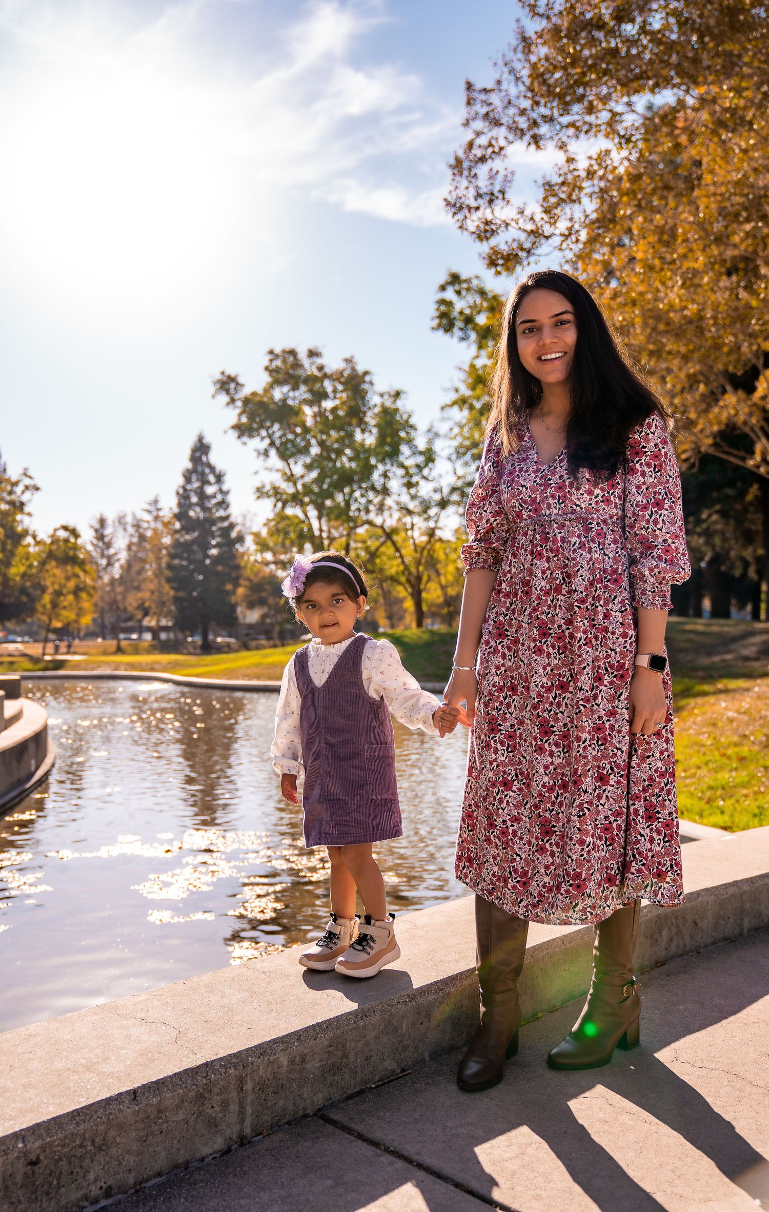 Two people in fall clothing stand by a pond on a sunny autumn day with golden trees in the background.
