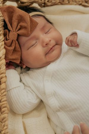 Sleeping baby wearing a white outfit and brown bow headband in a basket.