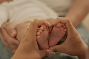 Tiny newborn baby feet cradled gently in caring hands on white bedding.