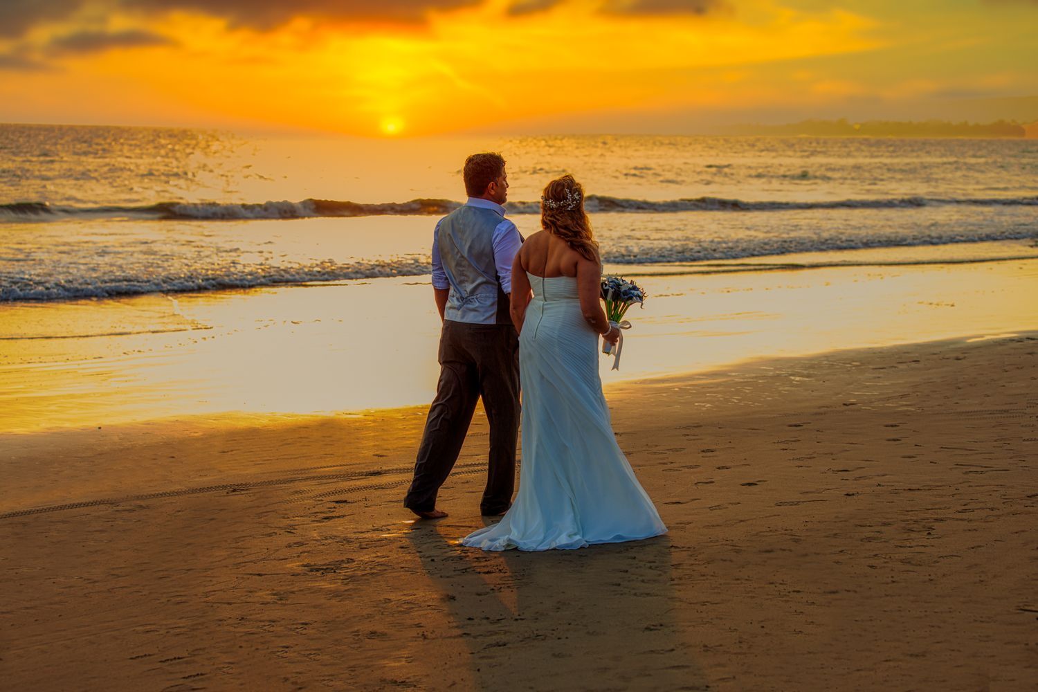 Newlywed couple standing on a beach at sunset, bride in white gown holding bouquet, groom in gray shirt.