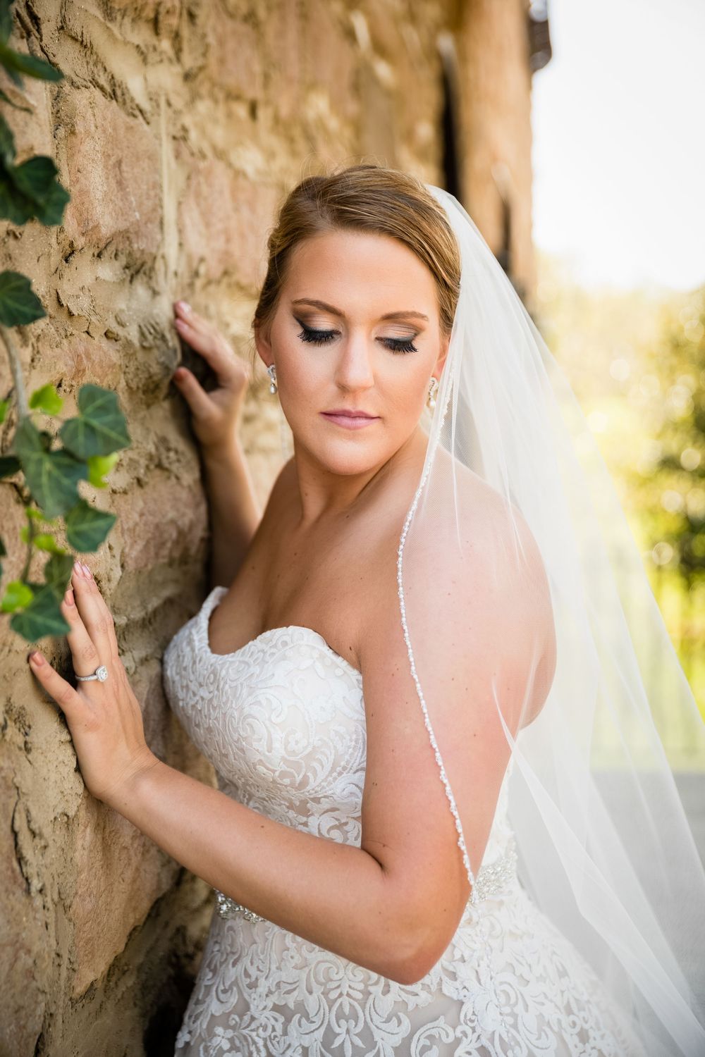 Bride in lace strapless gown and white veil leans against stone wall, looking down with elegant bridal makeup.