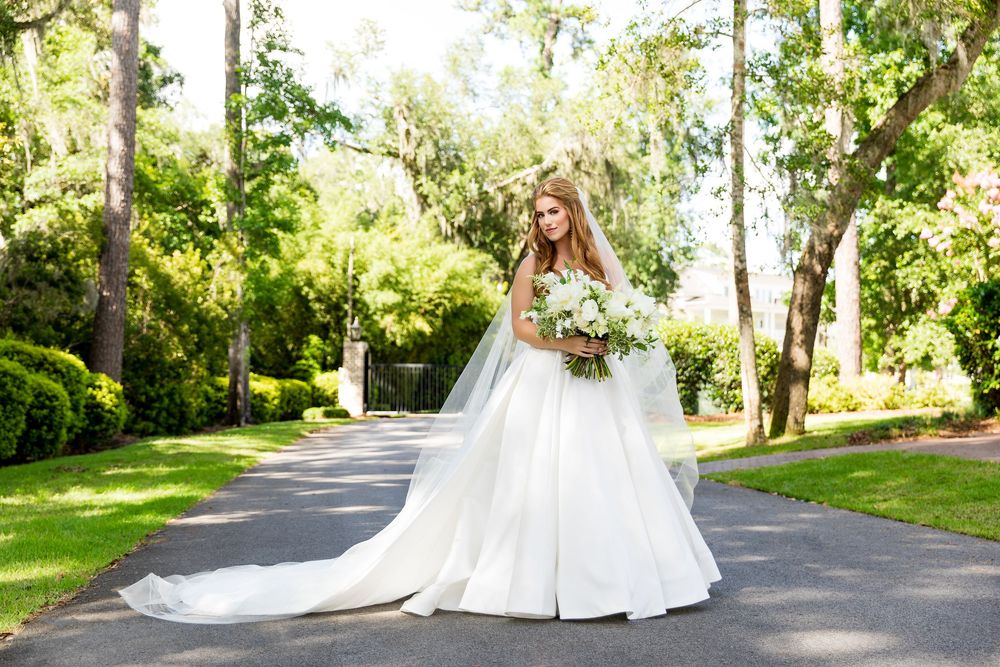 Bride in elegant white ball gown with long veil holding floral bouquet on tree-lined path outdoors.