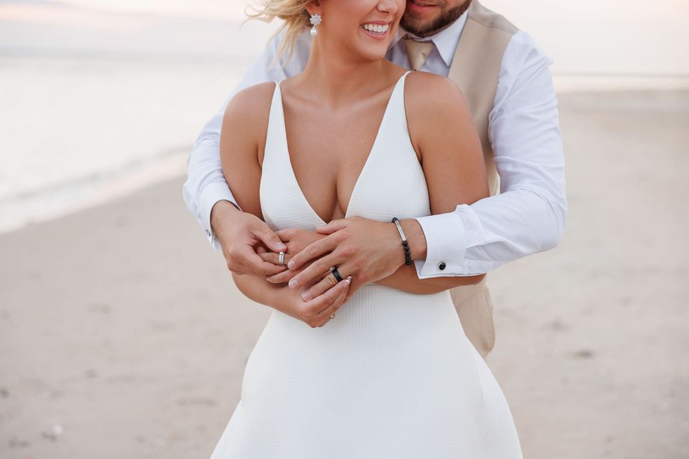 Couple embracing on beach at sunset, bride in white V-neck gown showing engagement ring, groom in white shirt and tan vest.