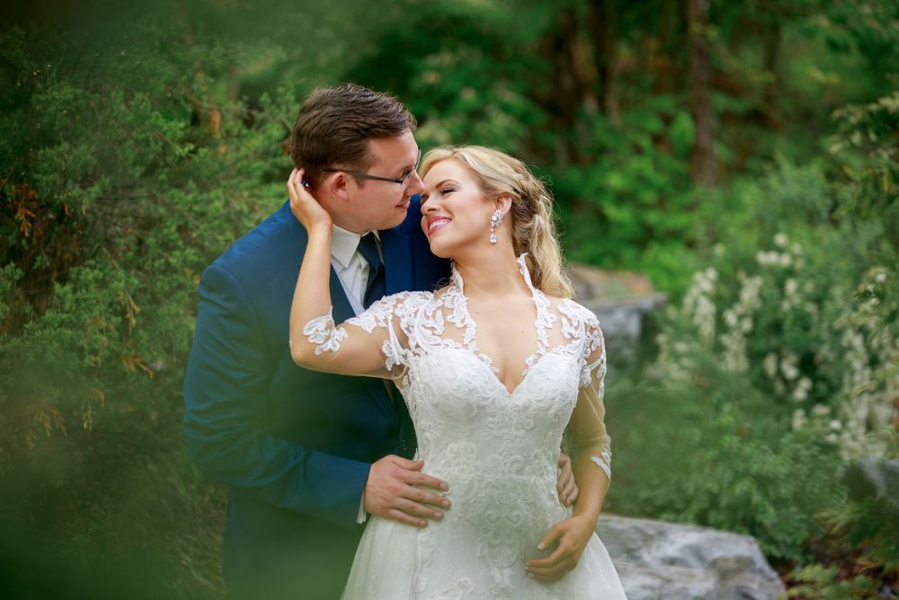 A groom in a blue suit kisses his smiling bride in a lace wedding dress outdoors among lush green trees.