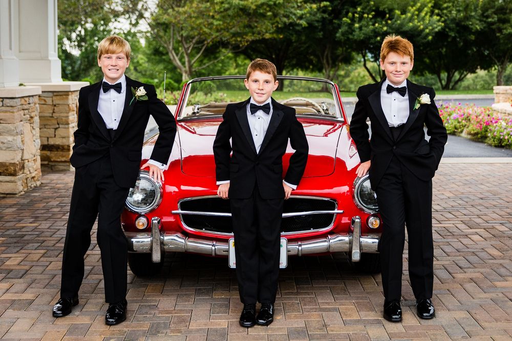 Three boys in black tuxedos with bow ties pose in front of a classic red convertible sports car.