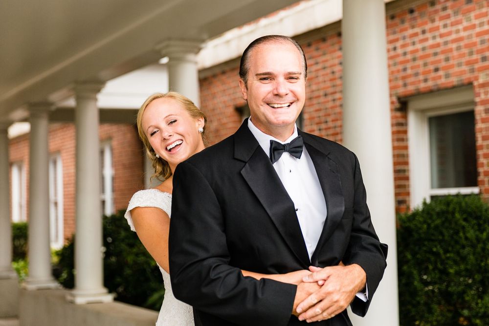 Smiling bride in white dress embraces groom in black tuxedo outside brick building with white columns.