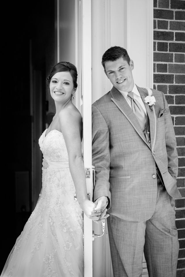 Black and white photo of a smiling bride and groom holding hands around a door on their wedding day.