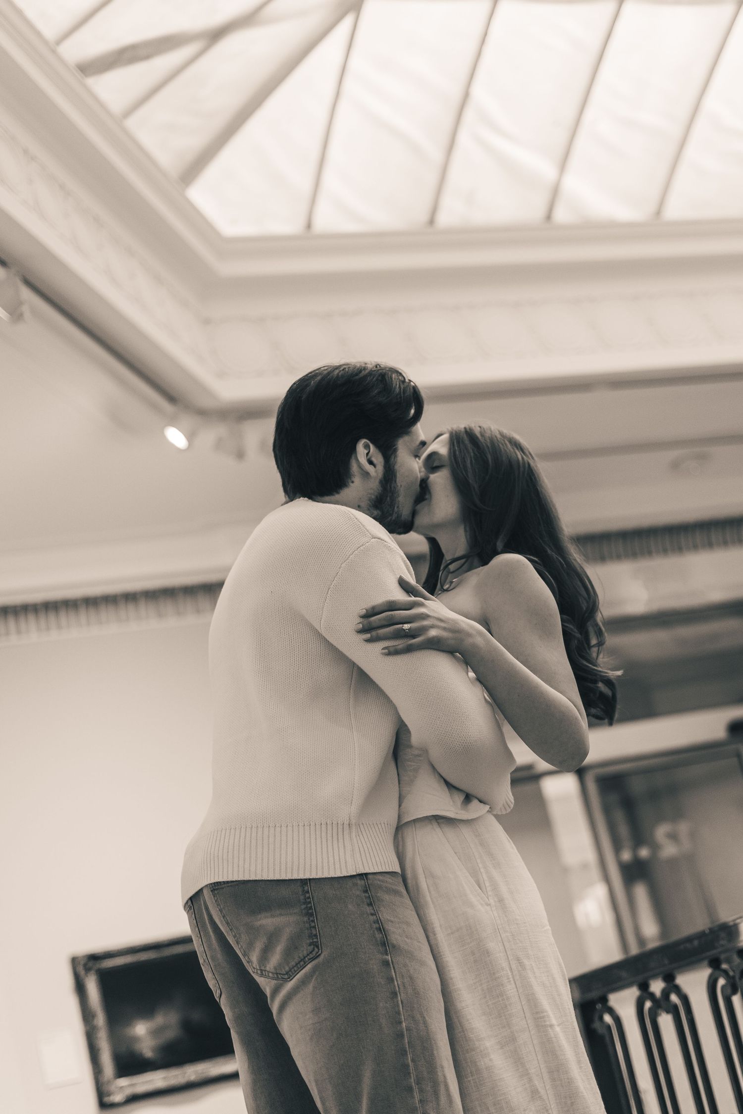 Black and white romantic photo of a couple sharing a tender kiss indoors beneath a skylight.