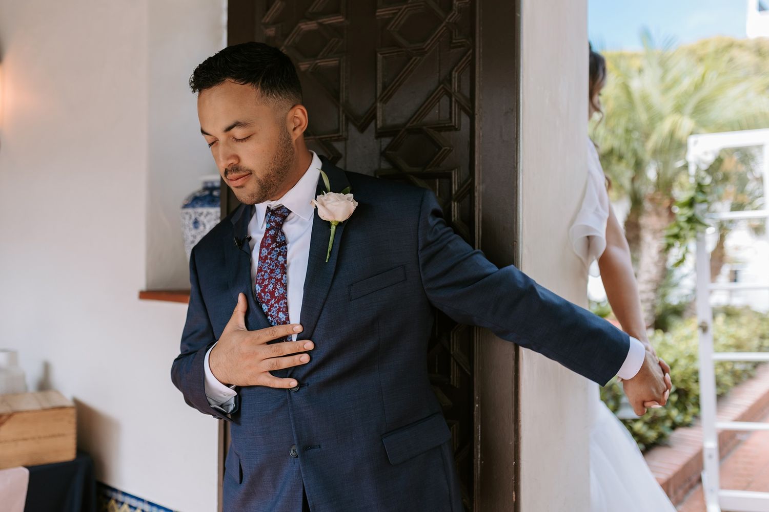 Groom in navy suit with floral tie and rose boutonniere holds the bride's hand before wedding ceremony.