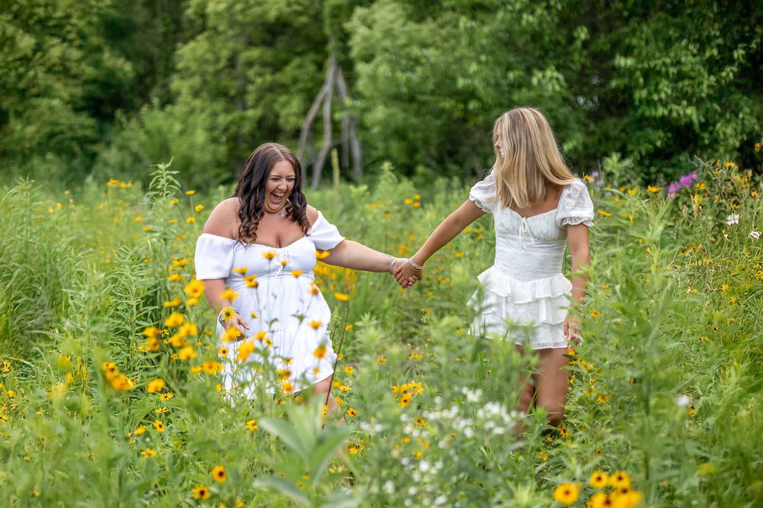 Two women in white dresses hold hands walking through a vibrant wildflower meadow with lush green trees in the background.