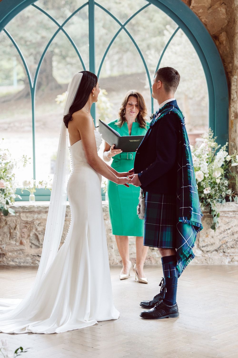 A wedding ceremony takes place under an arched window with the groom in traditional Scottish kilt and bride in white gown.