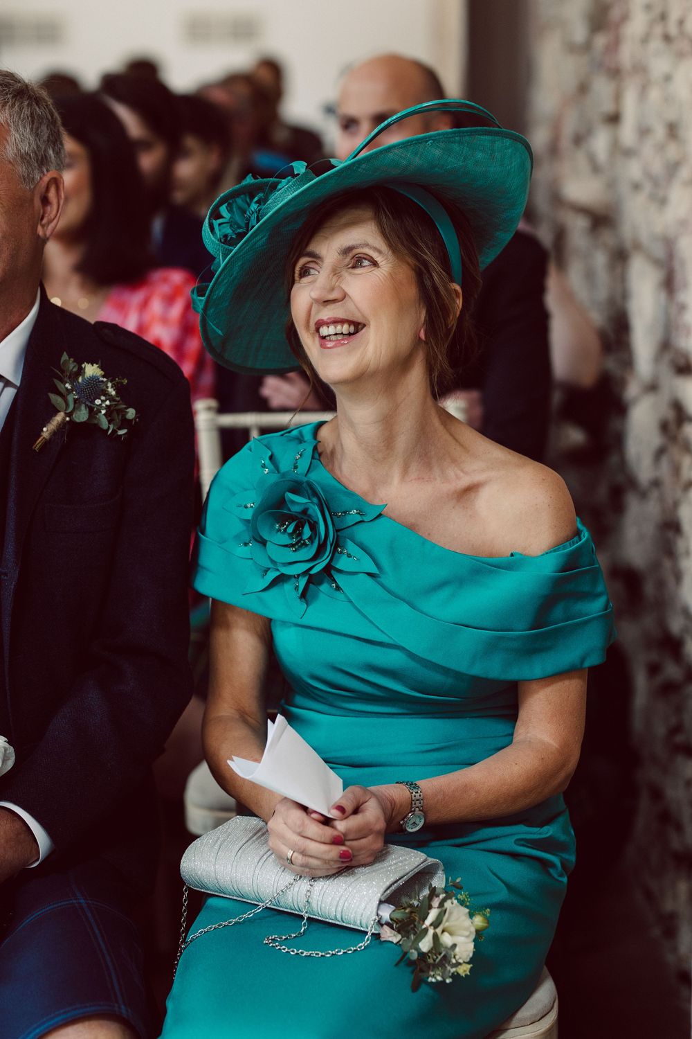 Wedding guest in elegant turquoise dress and matching wide-brimmed hat smiles during celebration.