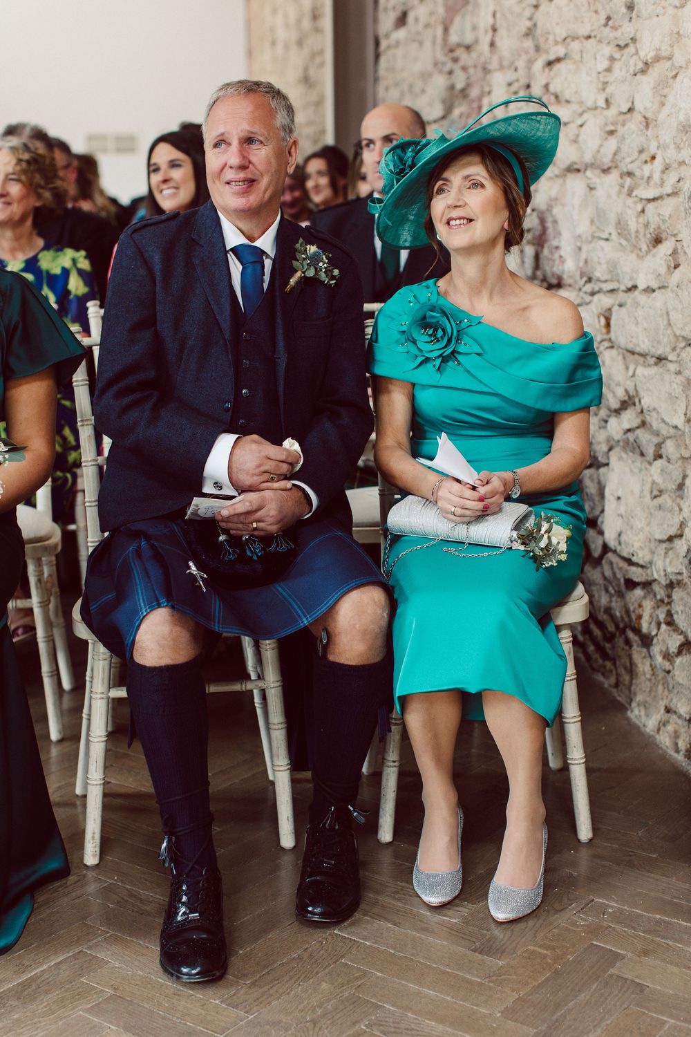 Wedding guests in turquoise attire sit in chairs against a stone wall during an outdoor ceremony.