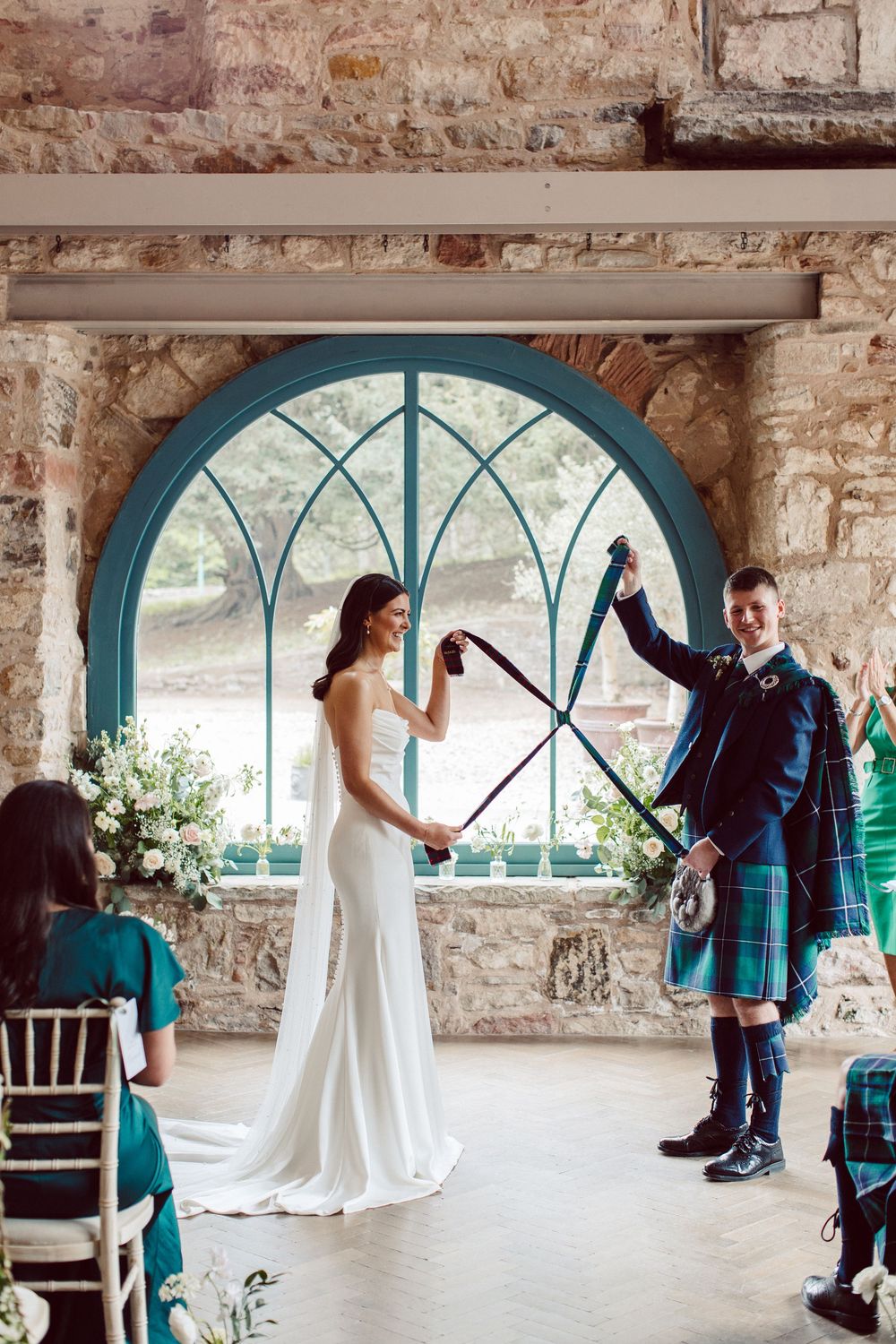 Wedding couple in traditional Scottish attire dancing together in a rustic stone venue with arched windows.