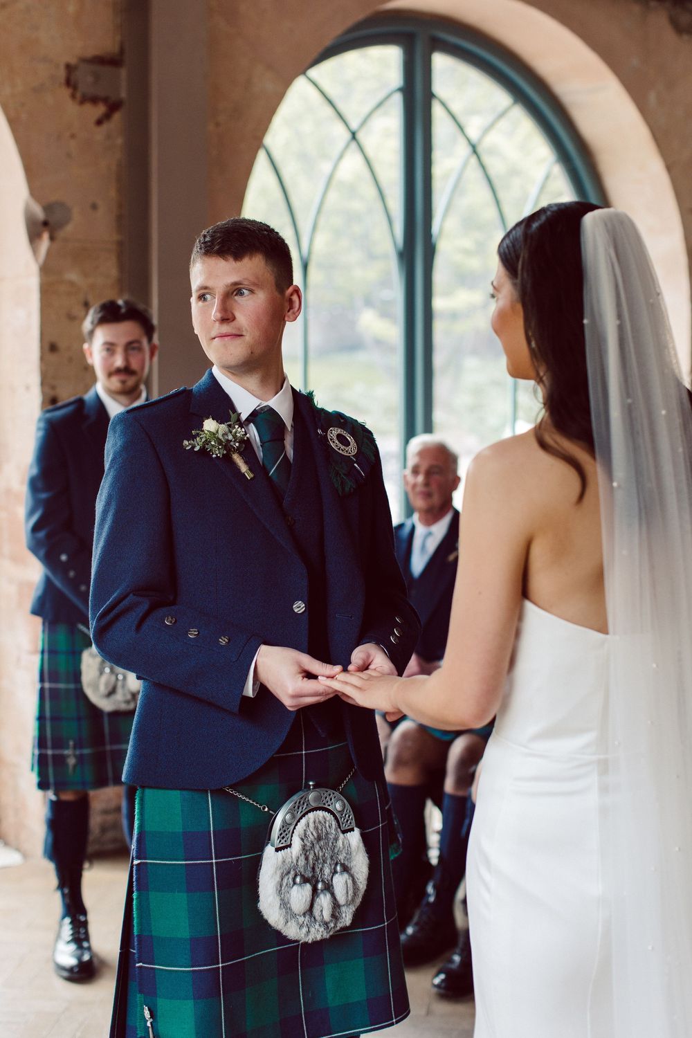 Wedding ceremony with men in traditional Scottish kilts and formal attire gathered in an elegant arched venue.