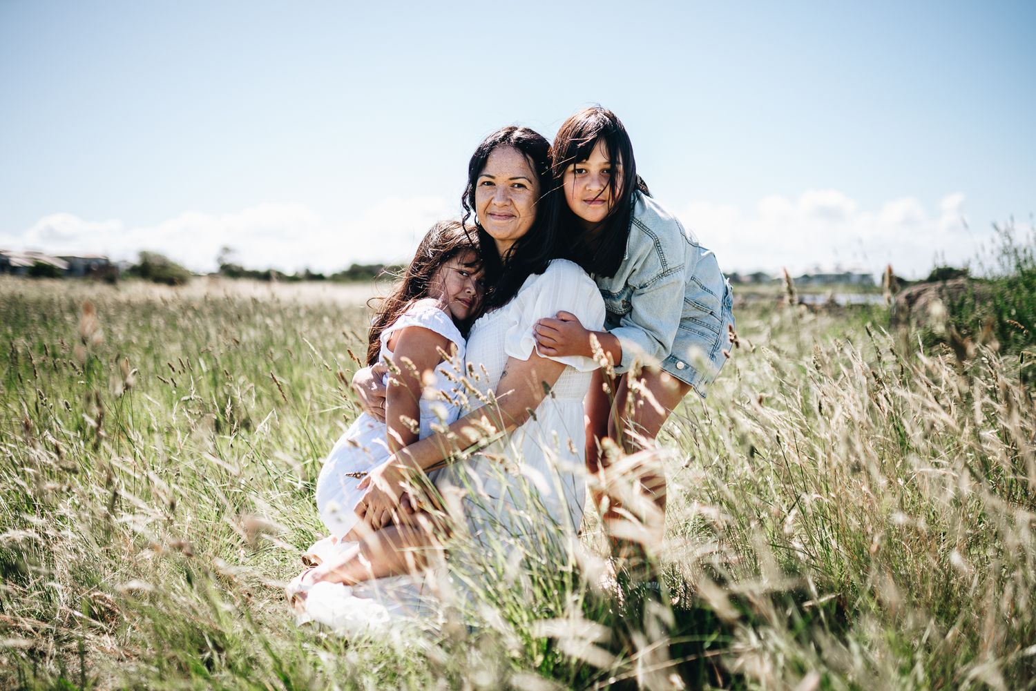 Mother and daughters embracing together in tall grass field on sunny day enjoying outdoor Melbourne Mother's Day photo session.