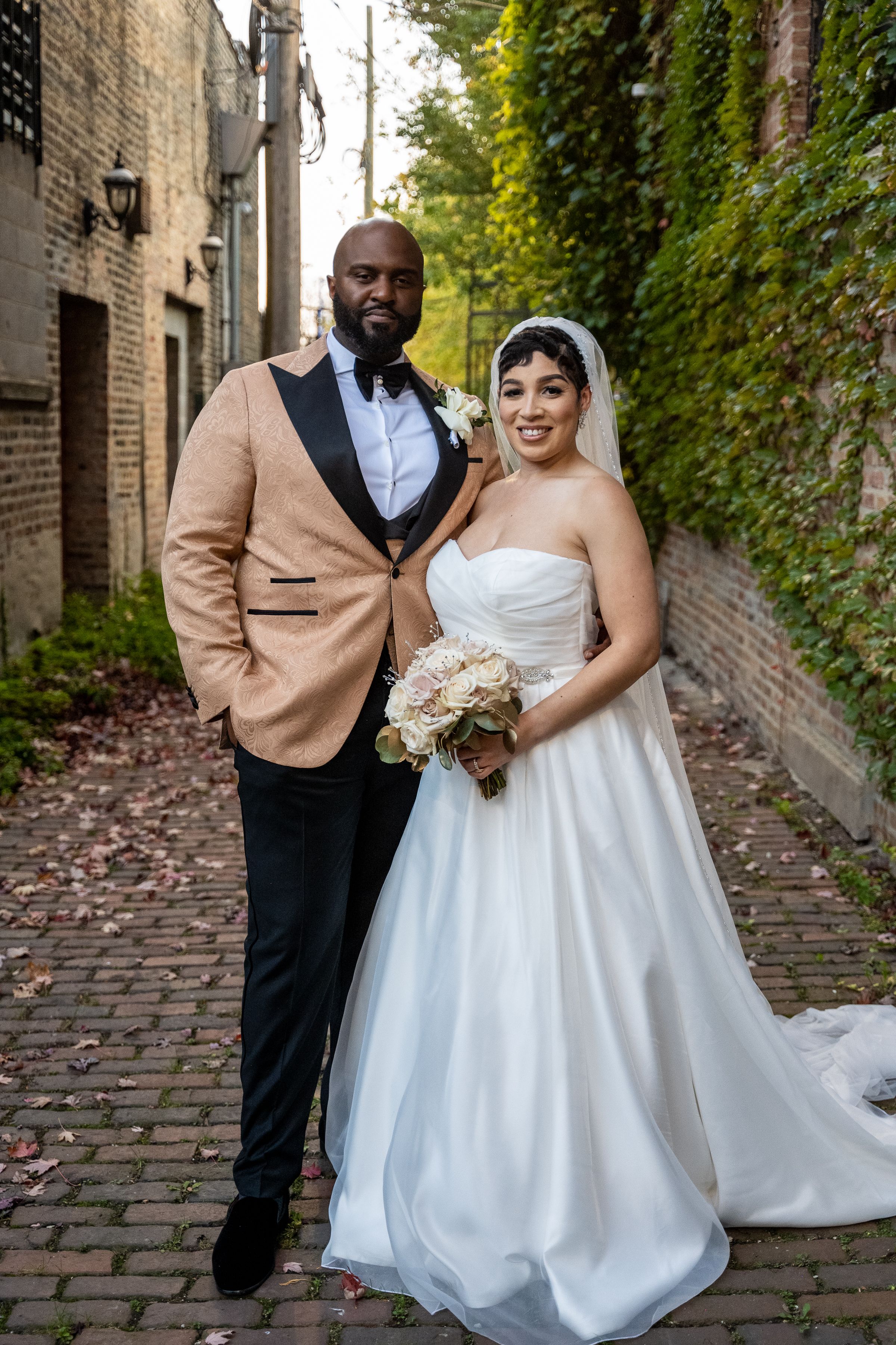A couple in wedding attire pose together on a charming cobblestone alley with ivy-covered brick walls.