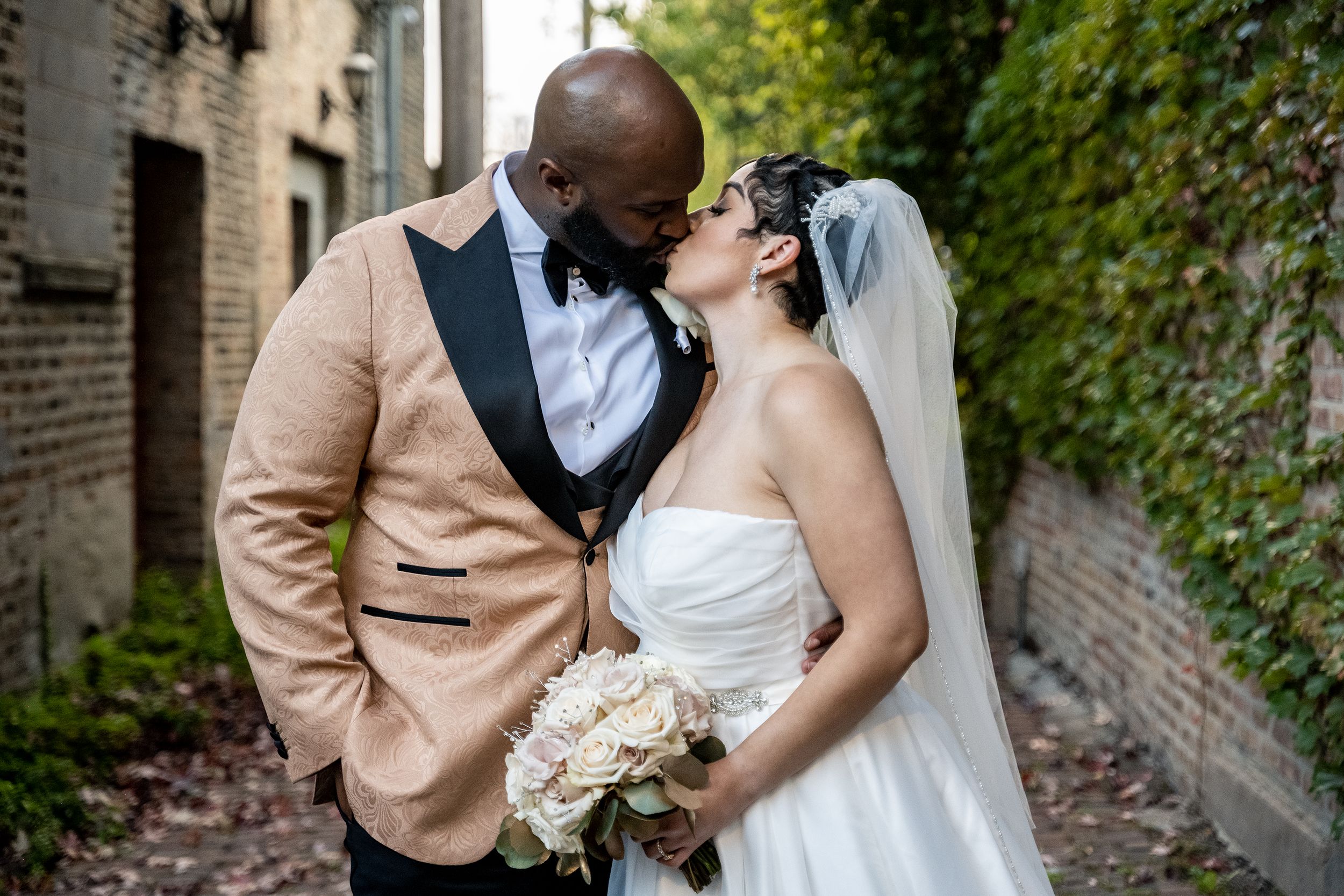 A wedding couple embraces and kisses in front of a brick building with lush green ivy.