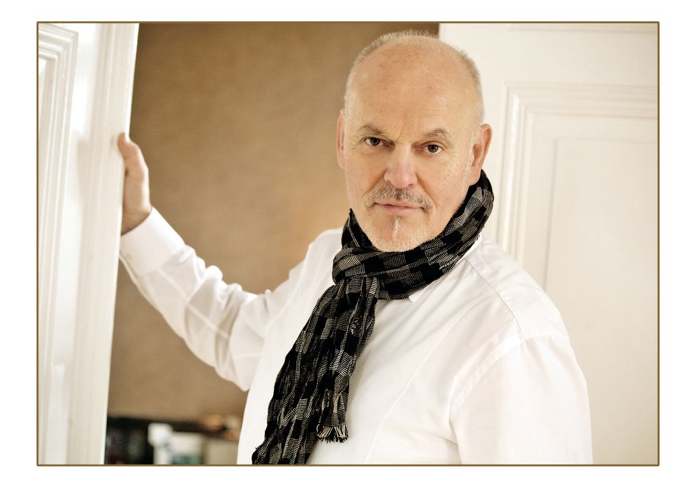 Middle-aged man in white shirt and dark scarf leaning against a white door frame in a stylish indoor setting.