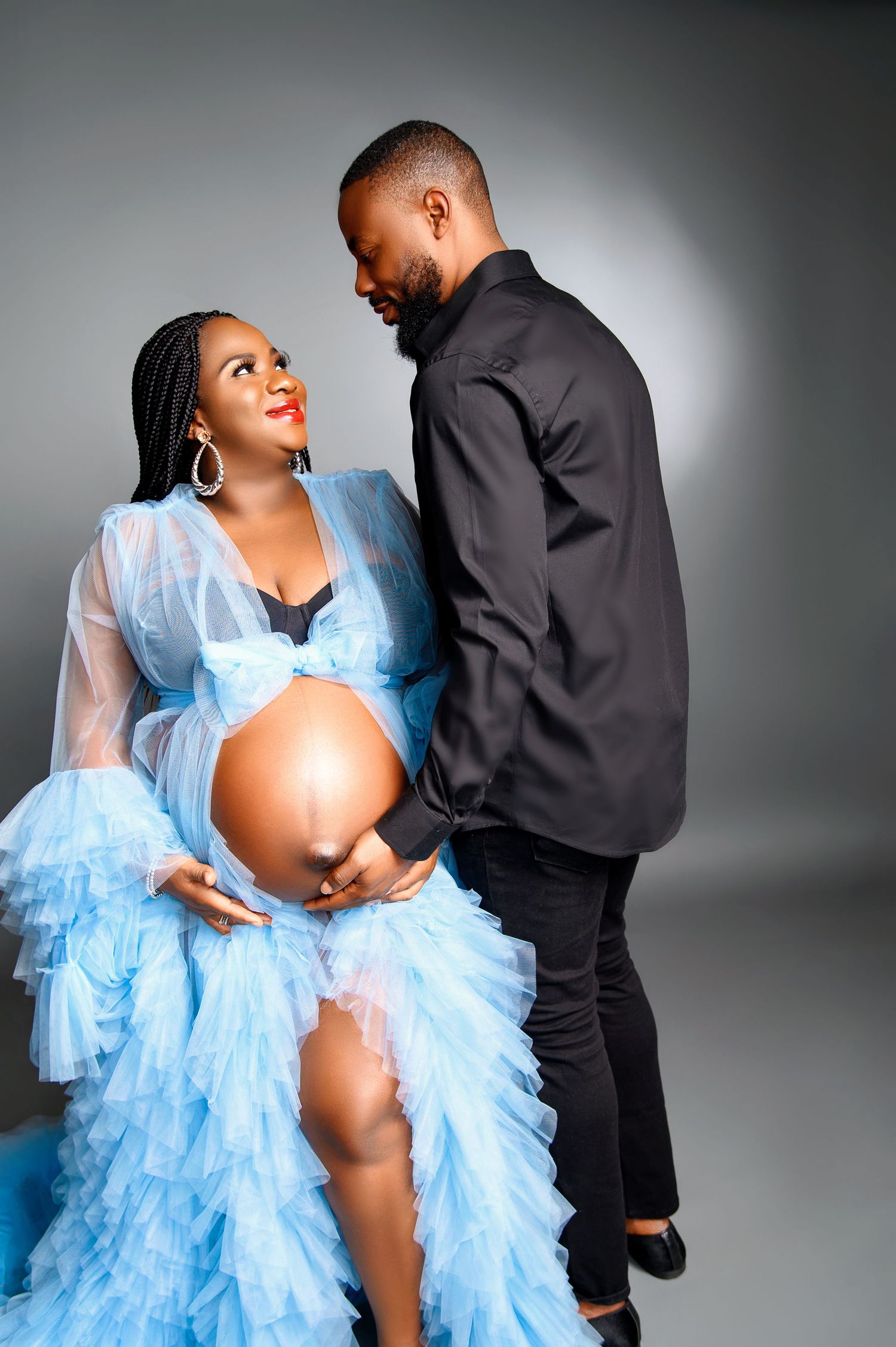 Maternity photoshoot featuring a couple in a blue tulle gown and black outfit against a gray backdrop.