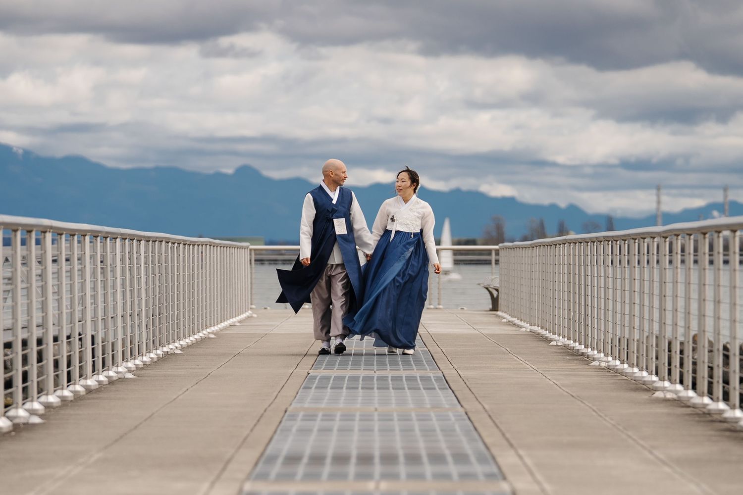 People sit on a long wooden pier overlooking a scenic mountain landscape under dramatic cloudy skies.