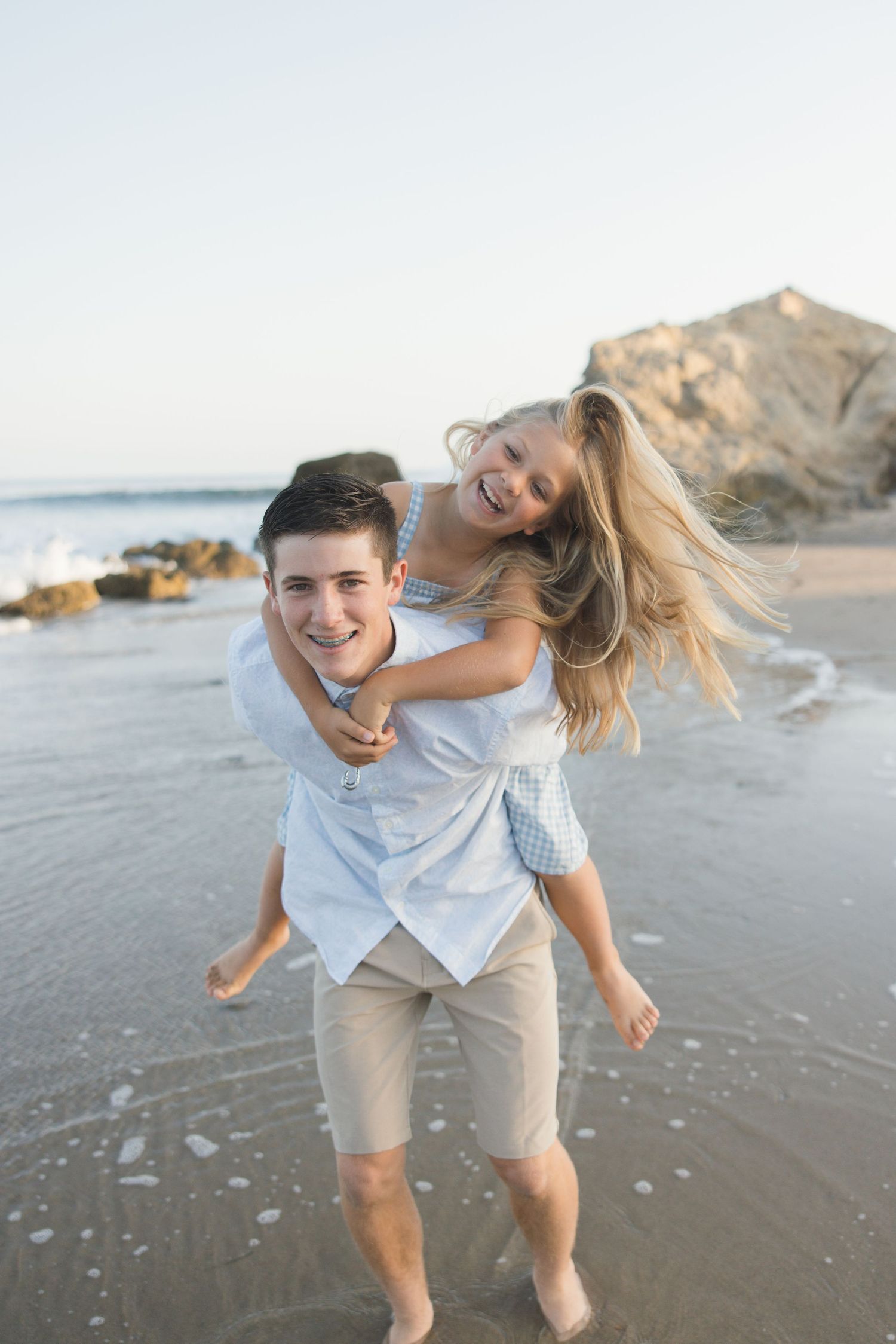 Family session at Leo Carrillo Beach in Malibu with an older brother giving his sister a piggyback ride in a light and airy coastal setting
