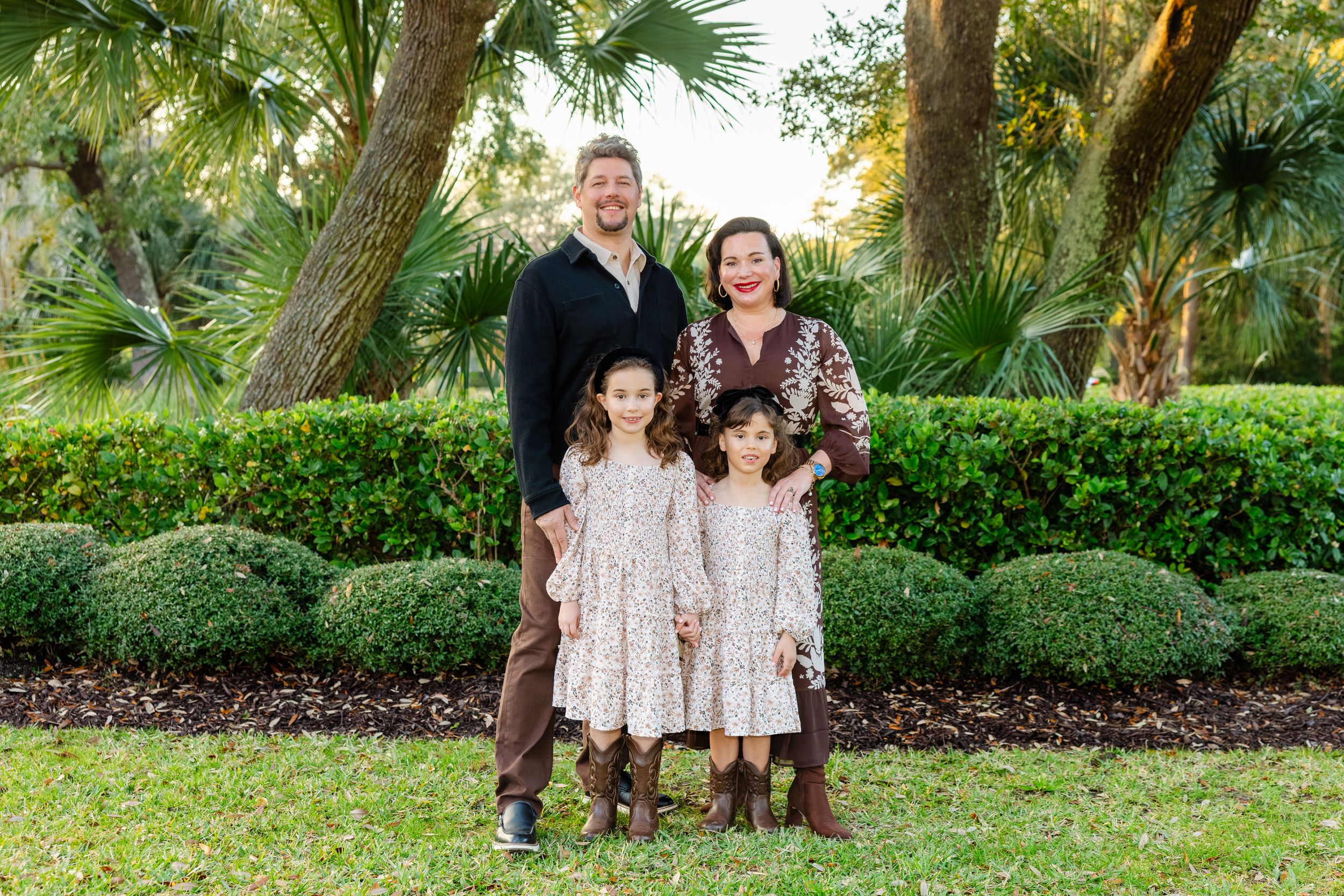 Family of four with two young daughters in matching floral dresses posing in a sunny park.