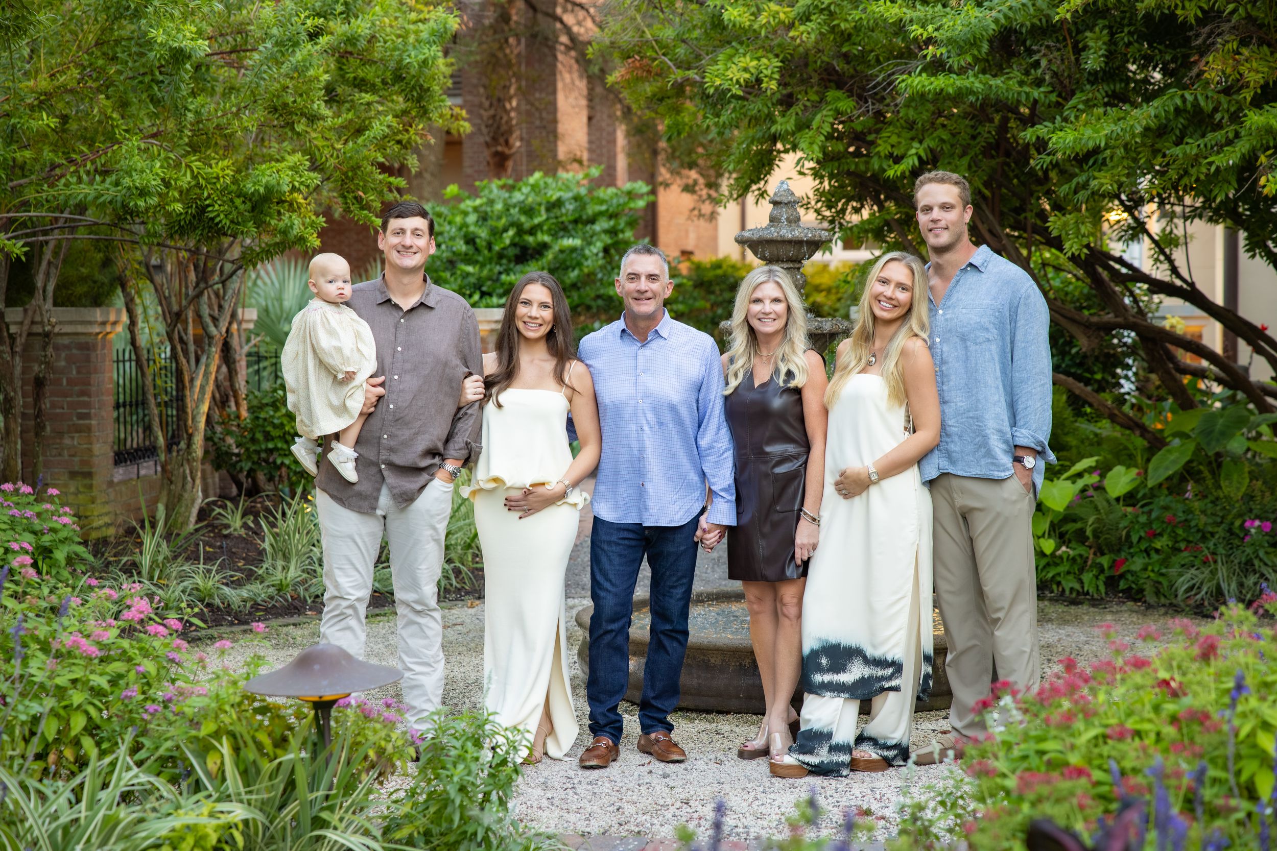 Family of seven adults and a baby posing together in a lush garden setting with greenery.