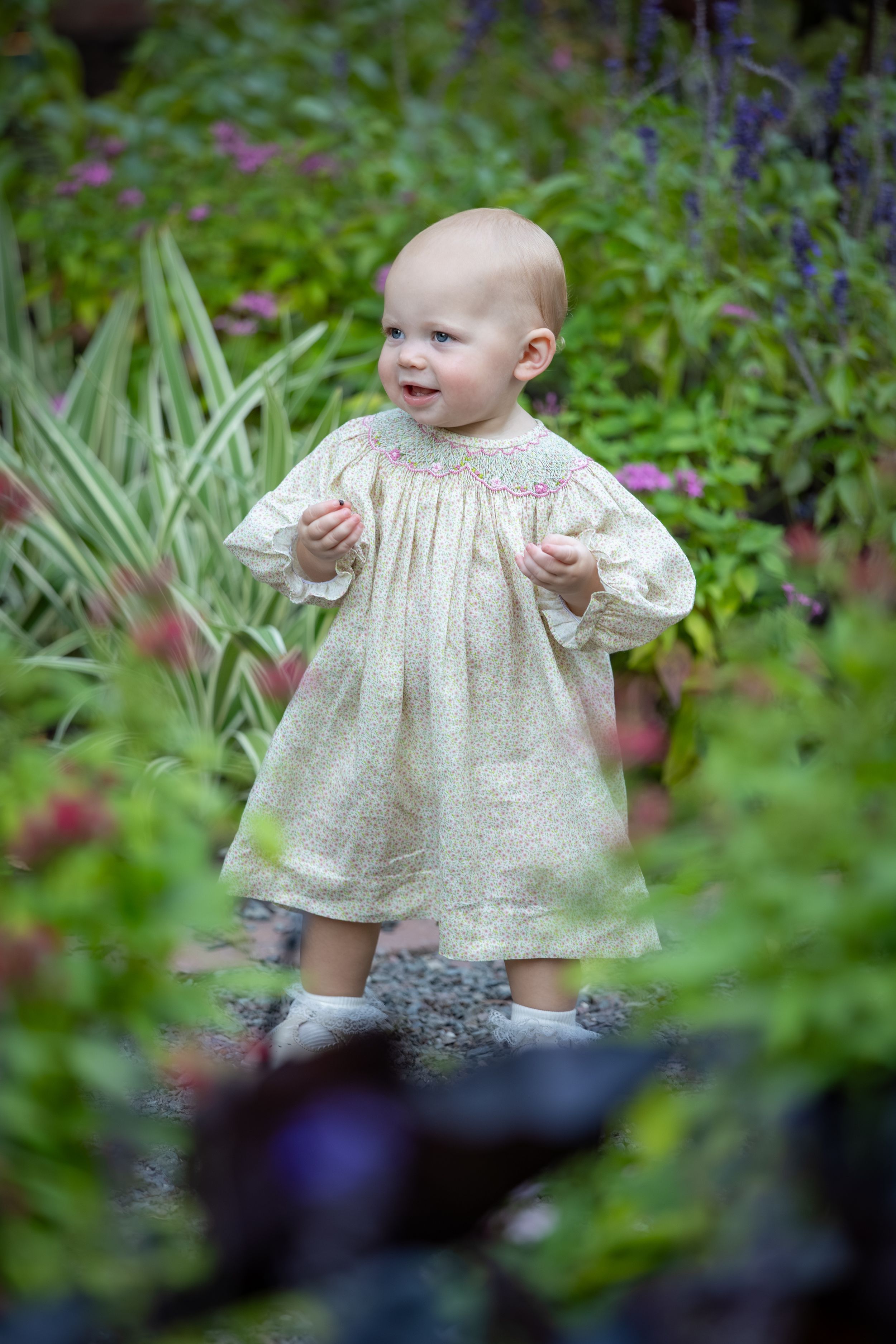 Adorable baby in cream smocked dress standing among colorful garden flowers and green foliage.