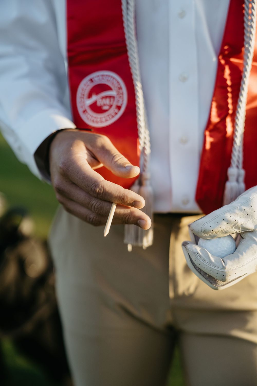 Graduate in white shirt with red sash and honor cords holding diploma outdoors at graduation ceremony.