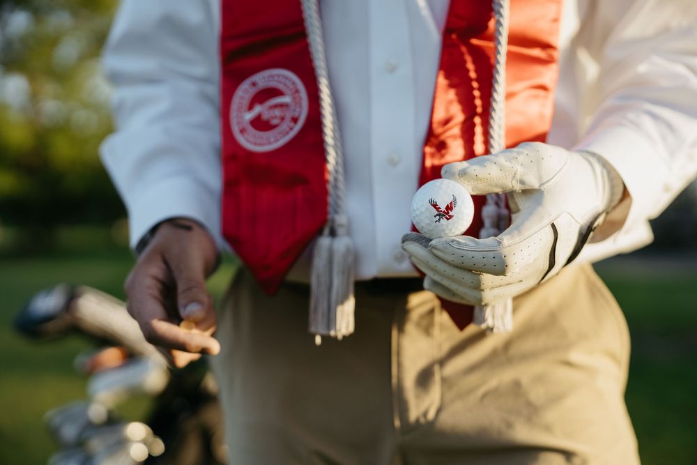 Golfer in graduation stole holding a golf ball and tee on a sunny golf course.